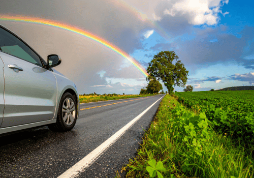 Car parked on a rural road with a bright rainbow overhead and shamrock graphics in the grass.