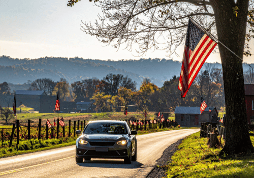 Start Summer Driving Season with Confidence in Your Vehicle Car driving on a rural road lined with American flags, with farms, trees, and hills in the background.